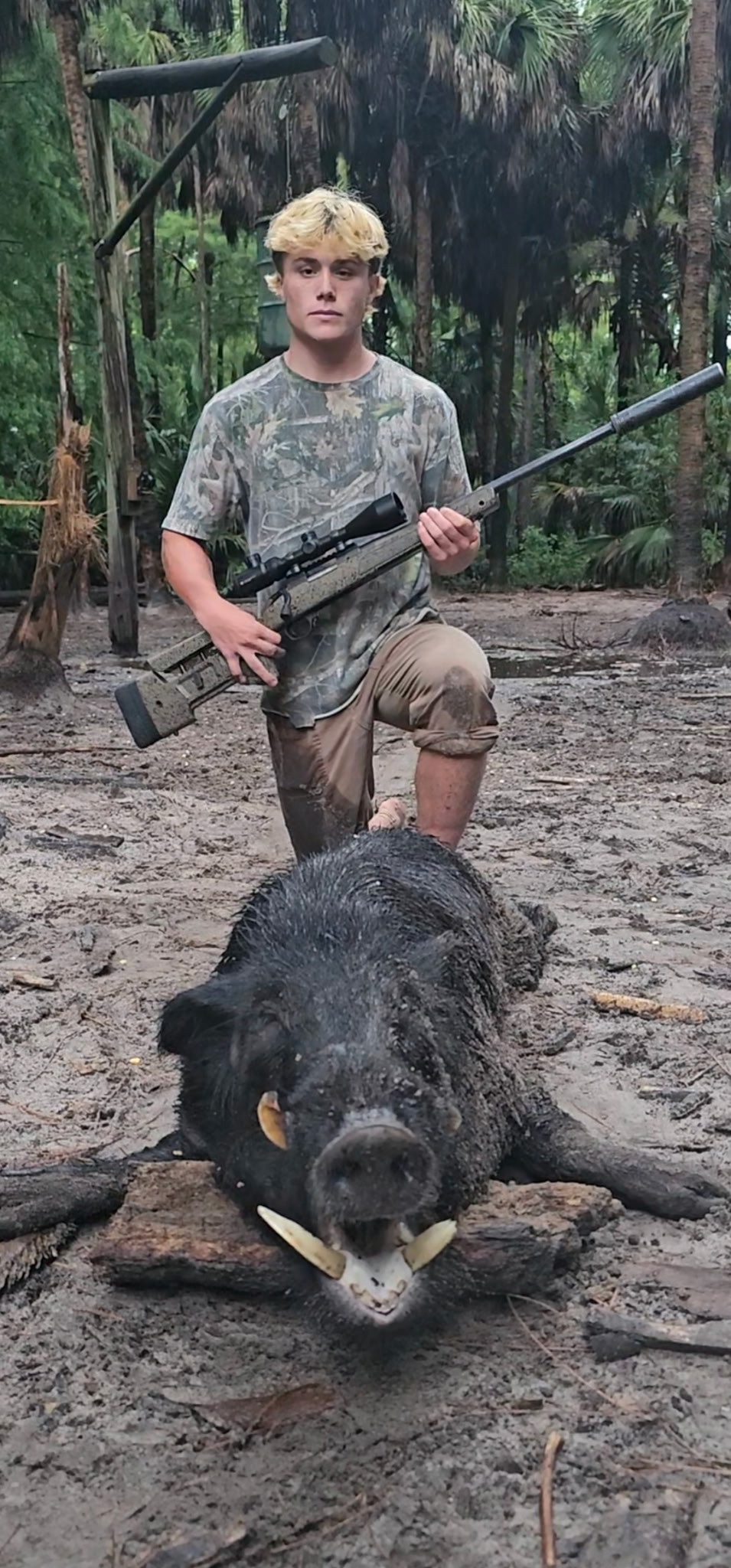 A young hunter holding a rifle beside a harvested wild boar in the woods