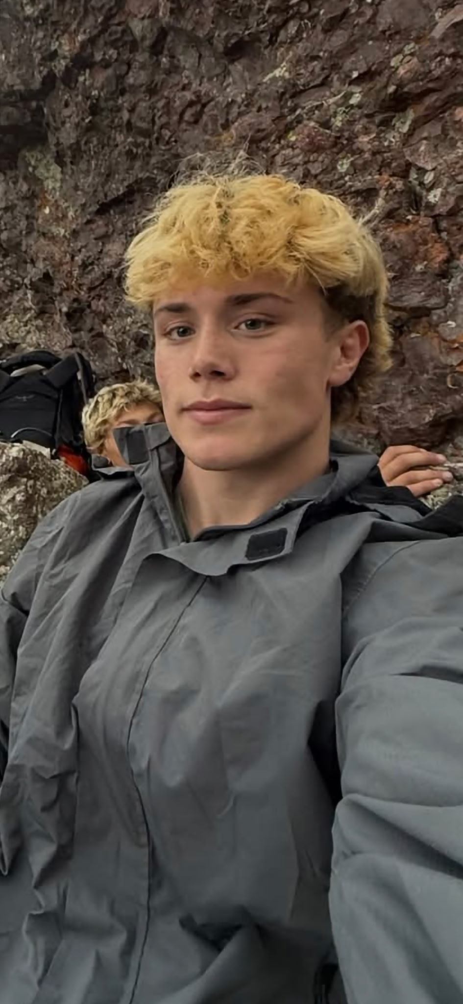 A young man in a gray hiking jacket among rocks, with companions behind him